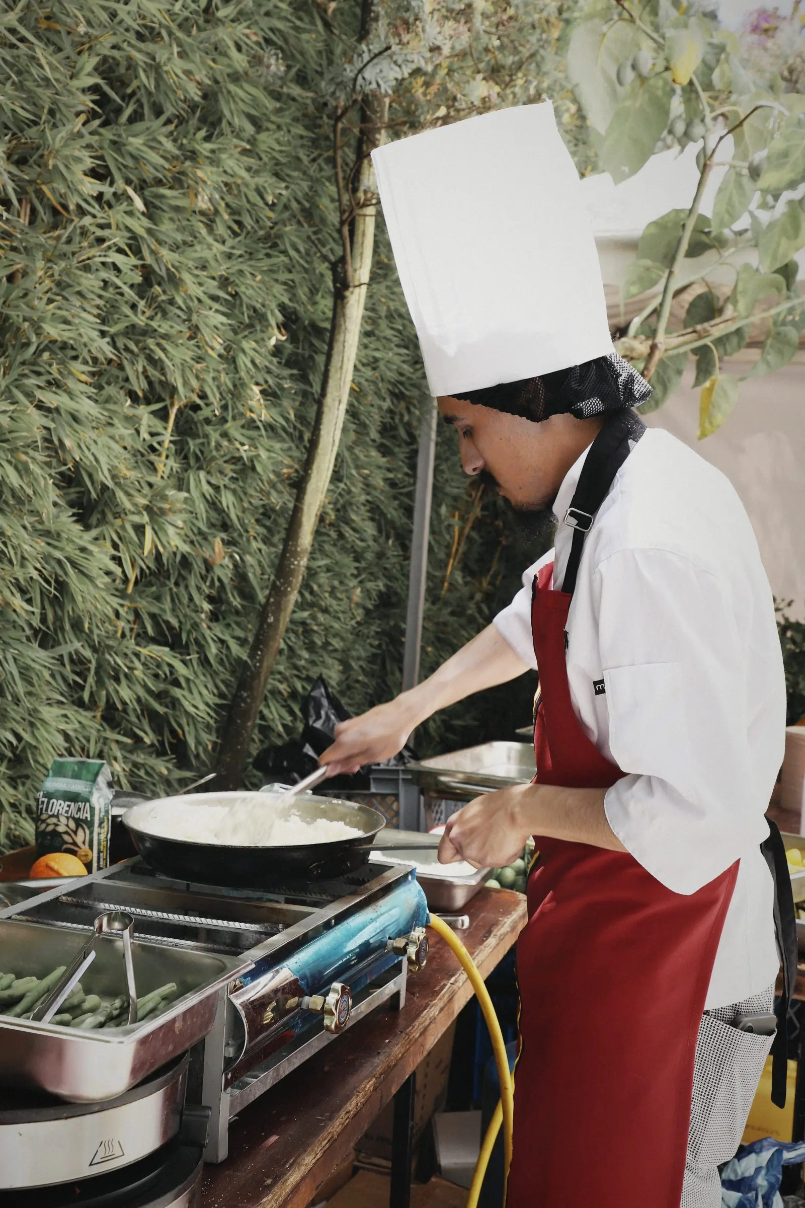 A cook in a tall white hat working at a field station behind the garden hedge.