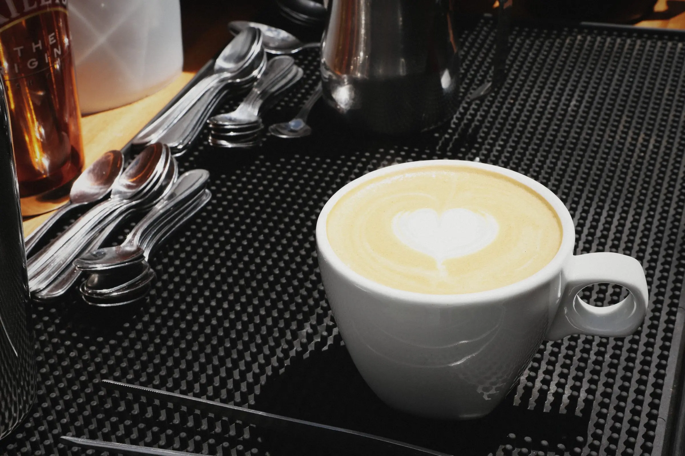 A cup of coffee with latte art on a black bar mat beside cutlery.